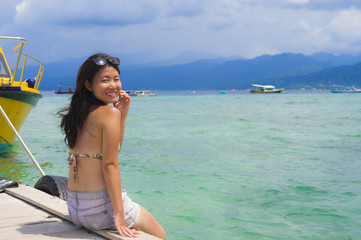 young Asian woman sitting happy on sea dock at Thailand beach looking at the horizon beautiful marine landscape with mountains enjoying holiday
