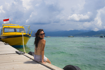 Obraz premium young Asian woman sitting happy on sea dock at Thailand beach looking at the horizon beautiful marine landscape with mountains enjoying holiday
