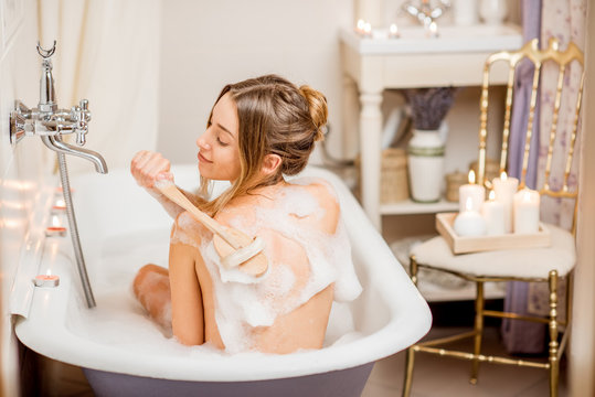 Young woman washing with brush in the beautiful vintage bath full of foam in the bathroom decorated with candles