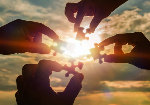 Collaborate four hands trying to connect a puzzle piece with a sunset background. A puzzle in hand against sunlight. One part of the whole. Symbol of association and communication. Business strategy.