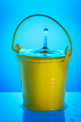 A small yellow bucket with water on a blue background. A drop of water falls into it. - 185015010