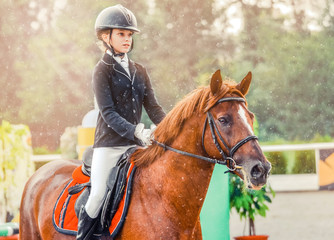 Young girl in uniform jumping with sorrel horse. Blond pretty little girl going jump a hurdle in a competition. Girl with red horse during equestrian showjumping.