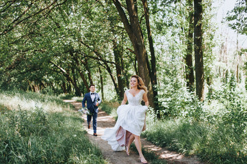 The bride and groom are happily running along the path in the forest. The bridegroom is catching up with the bride. The newlyweds laugh.