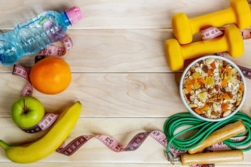 The concept of a way of life. Dumbbells, a plate with muesli, an orange, an apple, water, a skipping rope on a background of a wooden table.