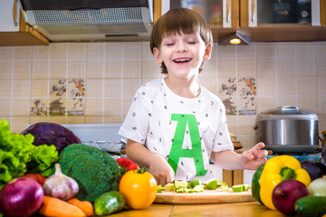 The young boy in cooking standing in the kitchen near table with vegetables