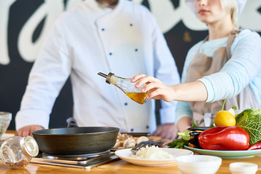 Close up view of young woman adding olive oil to gourmet dish while working with professional chef in restaurant kitchen