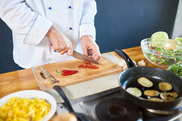 Close up of  unrecognizable chef working in kitchen, cutting red peppers and roasting eggplant on frying pan for gourmet culinary creation  in modern open kitchen