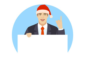 Businessman in Santa hat holding white blank poster and pointing up