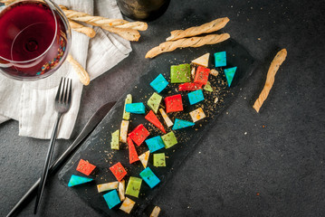 Trendy modern food. Multicolored colorful holland cheese - blue (lavender), red (hot pepper), green (basil), classic yellow cheese. With bread sticks, wine glass, black table. Copy space top view