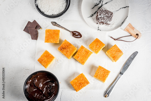 Australian cuisine. Preparation of traditional Australian dessert Lamington: biscuit in chocolate with coconut shaving powder. Top view. White table.