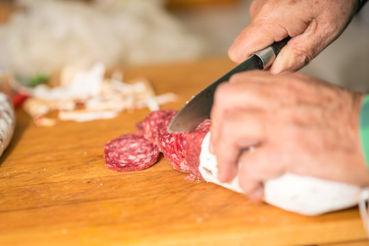Escena de comida donde destacan salchichones en barra y queso, junto con un cuchillo, listos para cortar o siendo cortados en rodajas o tacos. En una cocina dom&eacute;stica, en Madrid, Espa&ntilde;a