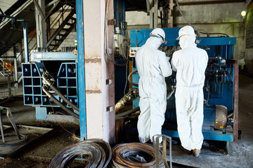 Back view portrait of two factory workers wearing biohazard suits operating machine on modern industrial plant, copy space