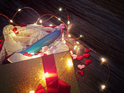 A blue plastic dildo vibrator is in an open gift box on a black wooden background. Nearby is a lid with a ribbon and a bow and scattered red hearts. Romantic background for sex shop