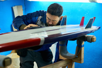 Portrait of mature worker carefully polishing surfing board with felt disk in yacht workshop, copy...