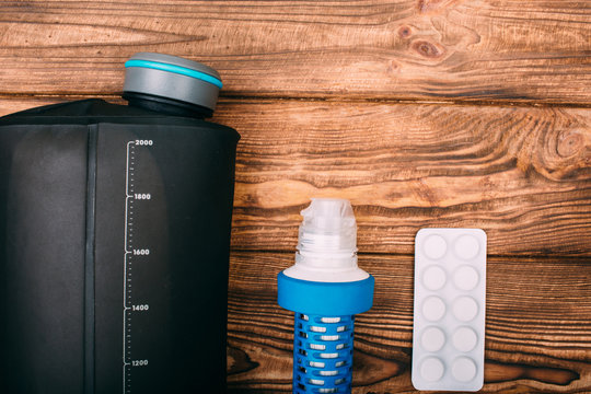 Kit For Water Purification During The Hiking. Items Include Filter For Water Purification, Tablets And A Bottle For Water On Wooden Background Top View