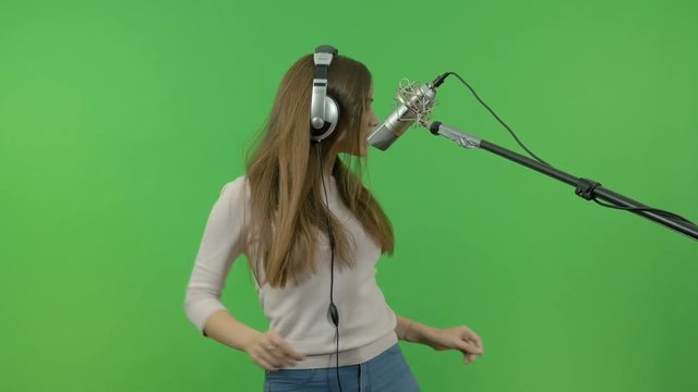 A singer with long hair sings into a studio microphone. On a green background.