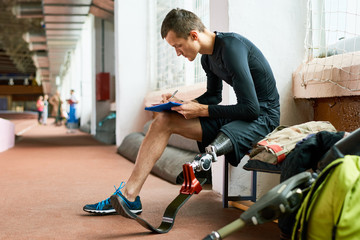 Young amputee athlete sitting on bench in gym and writing 