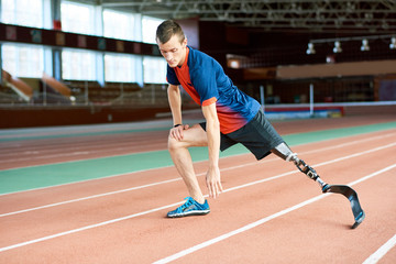 Full length portrait of young amputee athlete warming up before running practice in modern gym stretching legs on track, copy space