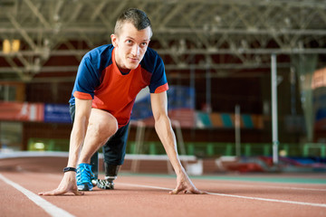 Motivational portrait of young amputee athlete on start position on running track in modern indoor...