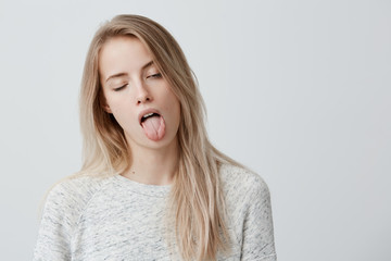Negative human reaction, feelings and attitude. Portrait of bored annoyed blonde girl in casual wear grimacing, sticking out her tongue, blinking, feeling nauseous because of bad smell or stink © Cookie Studio