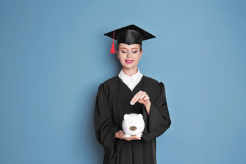 Young graduate putting coin into piggy bank on color background