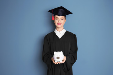 Young graduate holding piggy bank on color background