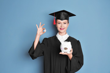 Young graduate holding piggy bank and coin on color background