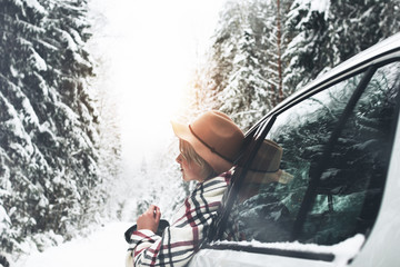 Boho style woman is sitting at the wheel of a car and look into the great distance. Pretty girl hipster in scarf and hat enjoying winter snow forest road. Flare effect