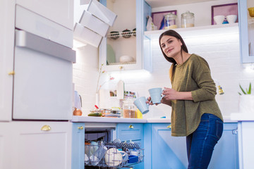 Side view of the young woman who standing in the kitchen and holding the cups in hands