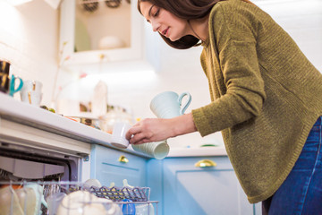 Side view of the young woman who standing in the kitchen and holding the white cups