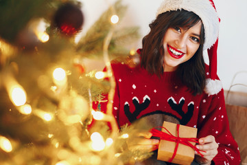 woman in santa hat holding stylish gift under christmas tree with garland lights and golden ornaments. space for text. seasonal greetings, happy holidays. merry christmas