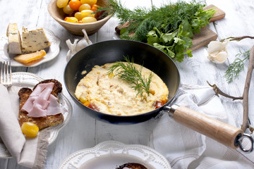 omelette with tomato in a frying pan for breakfast, cheese and greens on a white wooden background