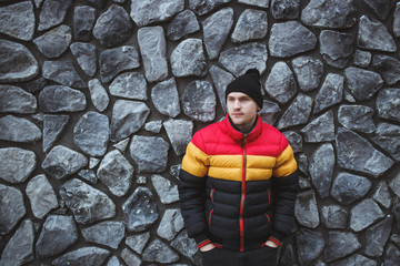 Handsome young man wearing colorful winter jacket and winter hat standing at stone gray wall outdoors. Winter time