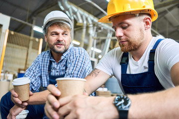 Portrait of two workers wearing hardhats taking break from work drinking coffee and resting sitting...