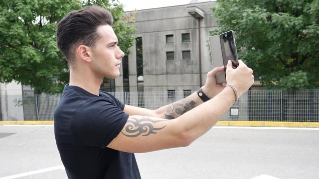 Young attractive man taking selfie photo outside in city street with tablet PC