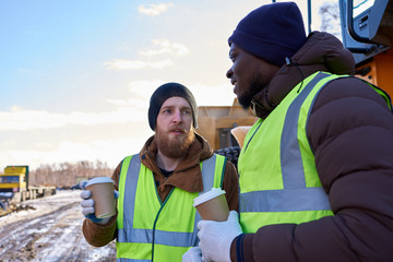 Portrait of two workers, one African-American, drinking coffee and chatting next to heavy...