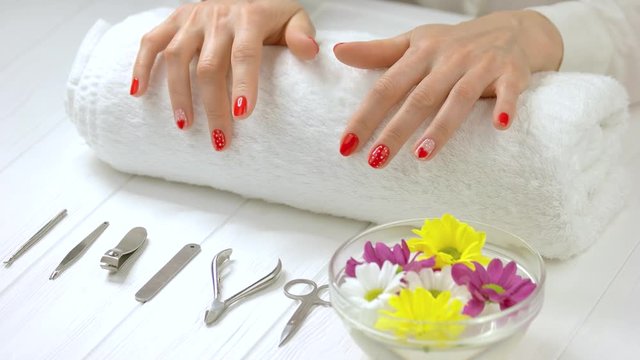 Hands with fresh manicure in spa salon. Well-groomed hands knocking on white towel with fingers. Manicure intrument, bowl with water and flowers. Spa treatment for hands.