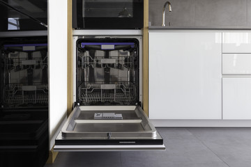 Opened new empty dishwasher in a modern loft kitchen in white, grey colors and wooden details. Incorporated dishwasher in the cabinets with hidden profile handle. 