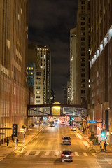 Street lights illuminate a city street with pedestrians and a skywalk in view