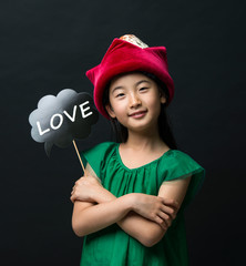 cute asian girl child dressed in a green dress holding a Christmas hat and a love stick on a black background.