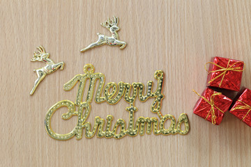 Gold Merry Christmas text and Red Gift Box on a brown wooden floor.