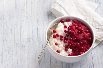 Rice pudding with cranberry jam and fresh cranberries in a white bowl on a light blue background, free space, selective focus. Delicious traditional breakfast