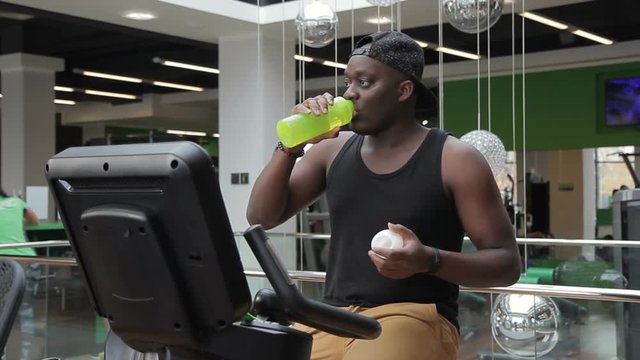 African American practices on an exercise bike and drinks water from bottle. Multiethical man trains leg muscles on sports equipment and takes sip from plastic container during break in gym.