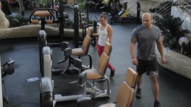 People go into gym and sit down on sports equipment for training. Man and woman go to fitness club and choose training machine to load muscles.