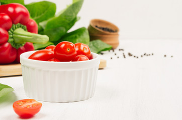 Ripe red cherry tomato in white bowl with fresh leaves salad on white wood board, closeup, copy space.