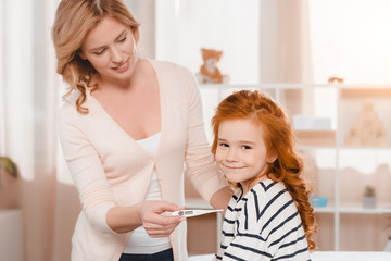 portrait of mother measuring daughters temperature with thermometer