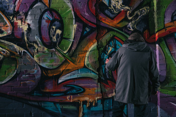 back view of street artist painting graffiti with aerosol paint on wall at night