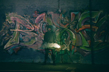 person holding smoke bomb and standing against wall with graffiti at night