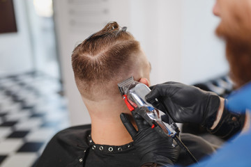 Close up of male professional hairdresser serving client by clipper. Ginger handsome stylish young man with big beard and short hair getting trendy haircut in black cape. Light white barber shop room.