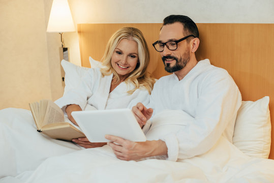 Happy Middle Aged Couple Reading Book And Using Digital Tablet While Lying Together In Bed In Hotel Room